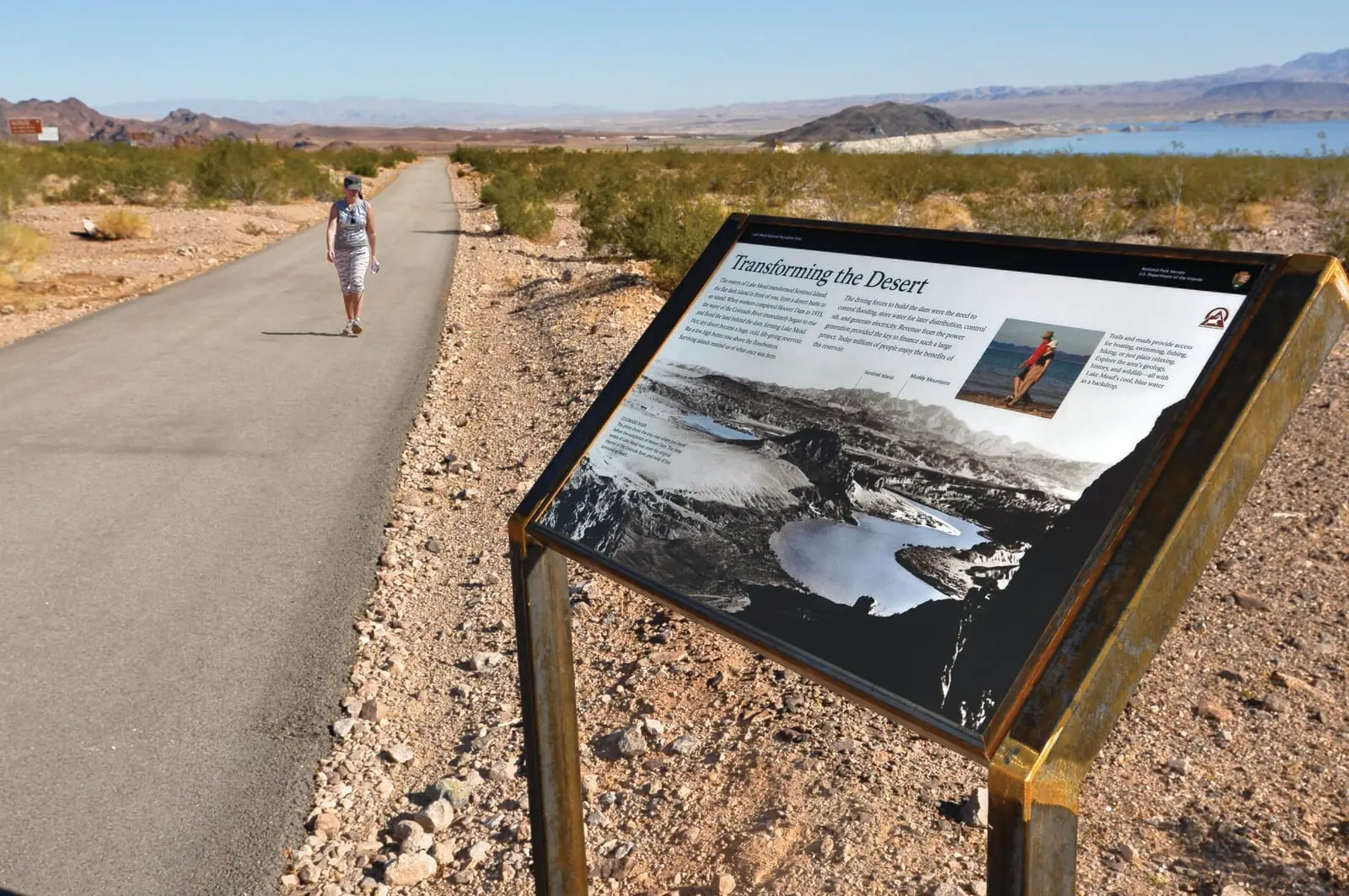 A walker on the paved River Mountains Loop Trail beside an interpretive sign, Lake Mead in the distance